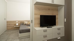 Un dormitorio con una cama, una mesita de noche con lámpara, un televisor sobre una estantería de madera y un armario con cajones.