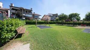 Un espacio al aire libre con césped, un conjunto de columpios de madera, un banco y un sendero pavimentado.
