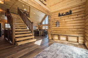 A log cabin interior with a wooden staircase, a fireplace, and a large window.