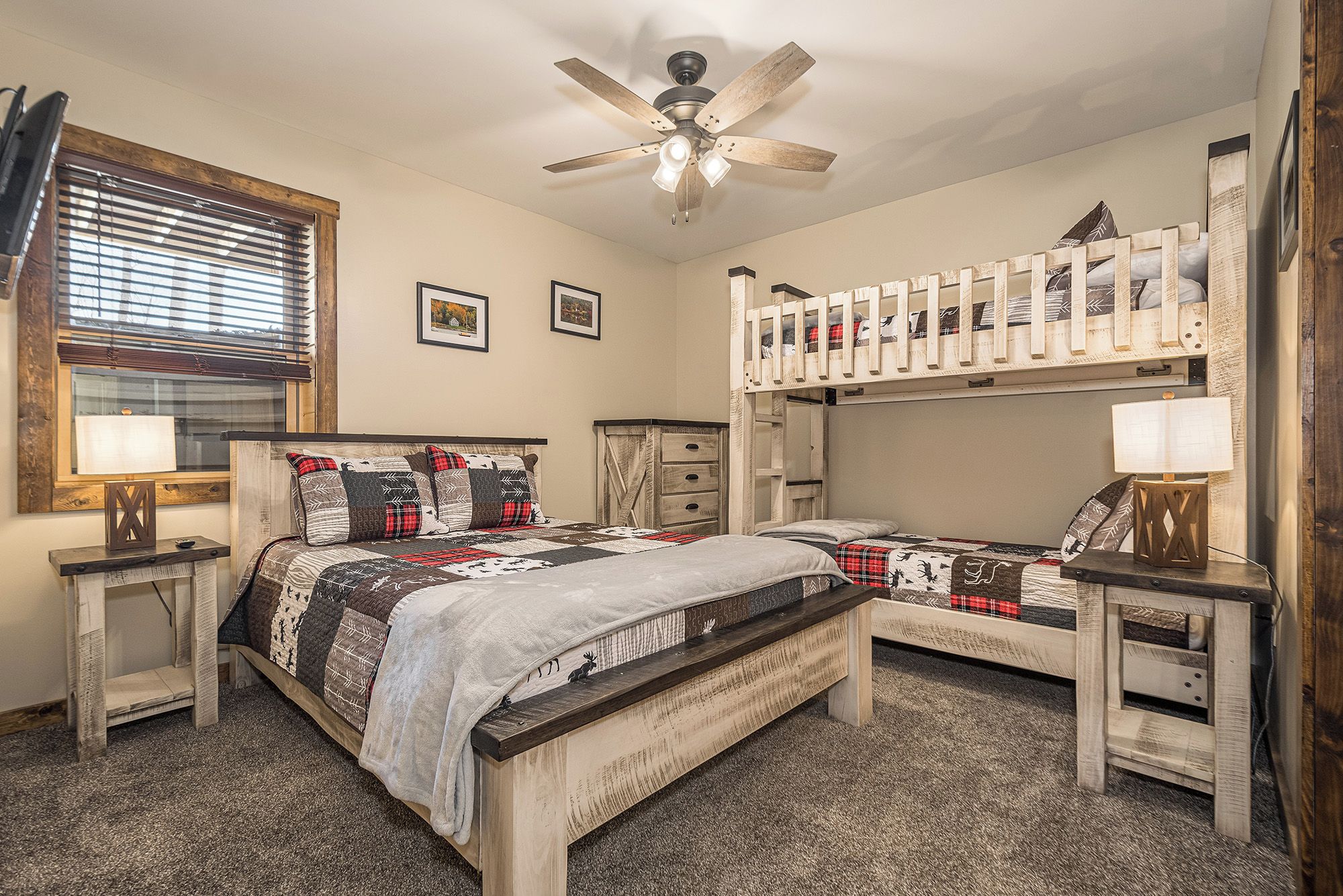 A bedroom with a bunk bed, a single bed, a ceiling fan, a window with blinds, and a bedside table with a lamp.