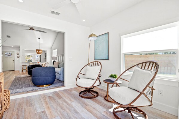 A modern living room with a large window, two wicker chairs, a blue ottoman, and a dining area in the background.