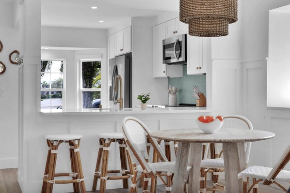 A modern kitchen with white cabinetry, stainless steel appliances, and a round dining table with wicker chairs. - The Blue Oasis - 3/2 POOL Home S Jax Beach (Jacksonville Beach)