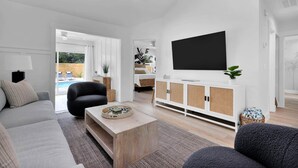Un salón moderno con sofá, una mesa de centro, un televisor sobre un mueble blanco y vistas al exterior.