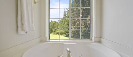 Un baño con una bañera grande, una ventana con vista a la vegetación y un perchero con dos toallas.