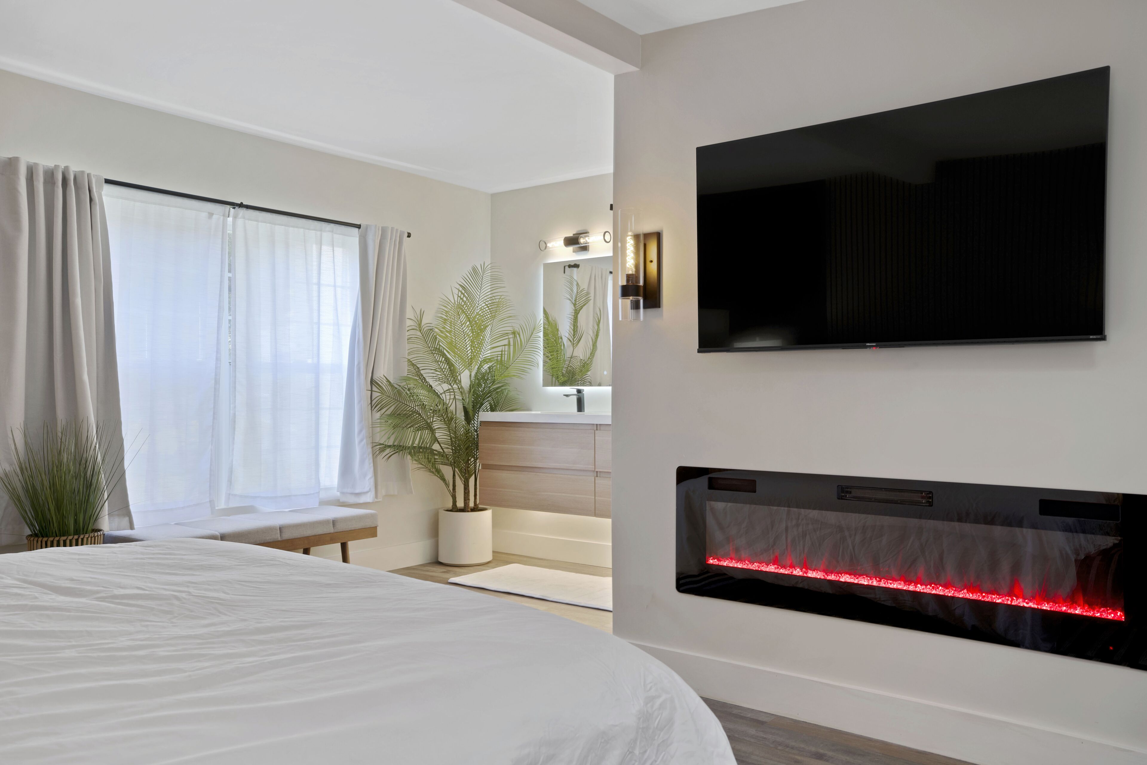 Un dormitorio moderno con cama, televisor montado, chimenea, ventana grande con cortinas y una planta en maceta.