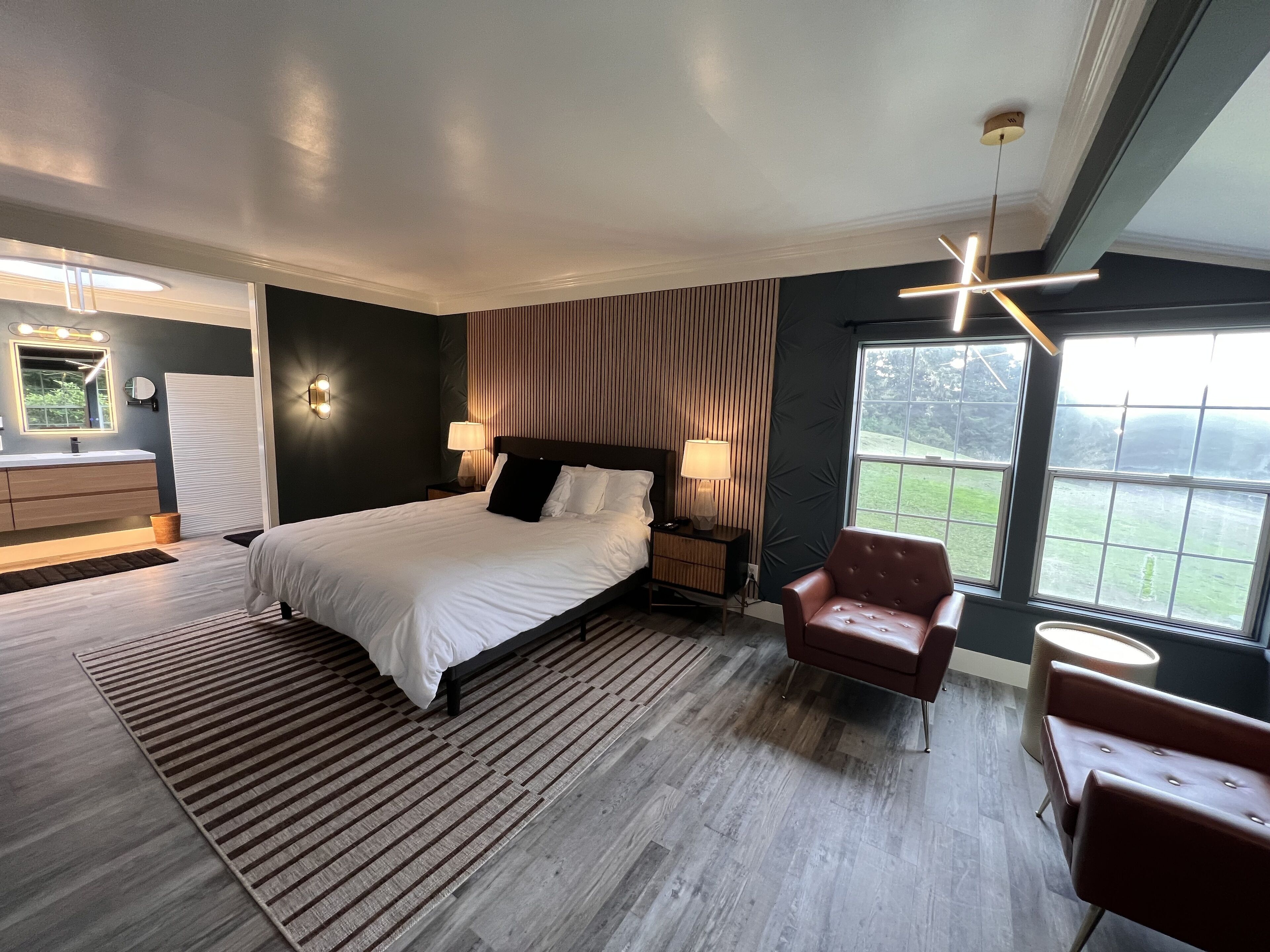 Une chambre moderne avec un grand lit, des lampes de chevet, un plancher en bois, un fauteuil rouge et une vue sur l’extérieur grâce à une grande fenêtre.