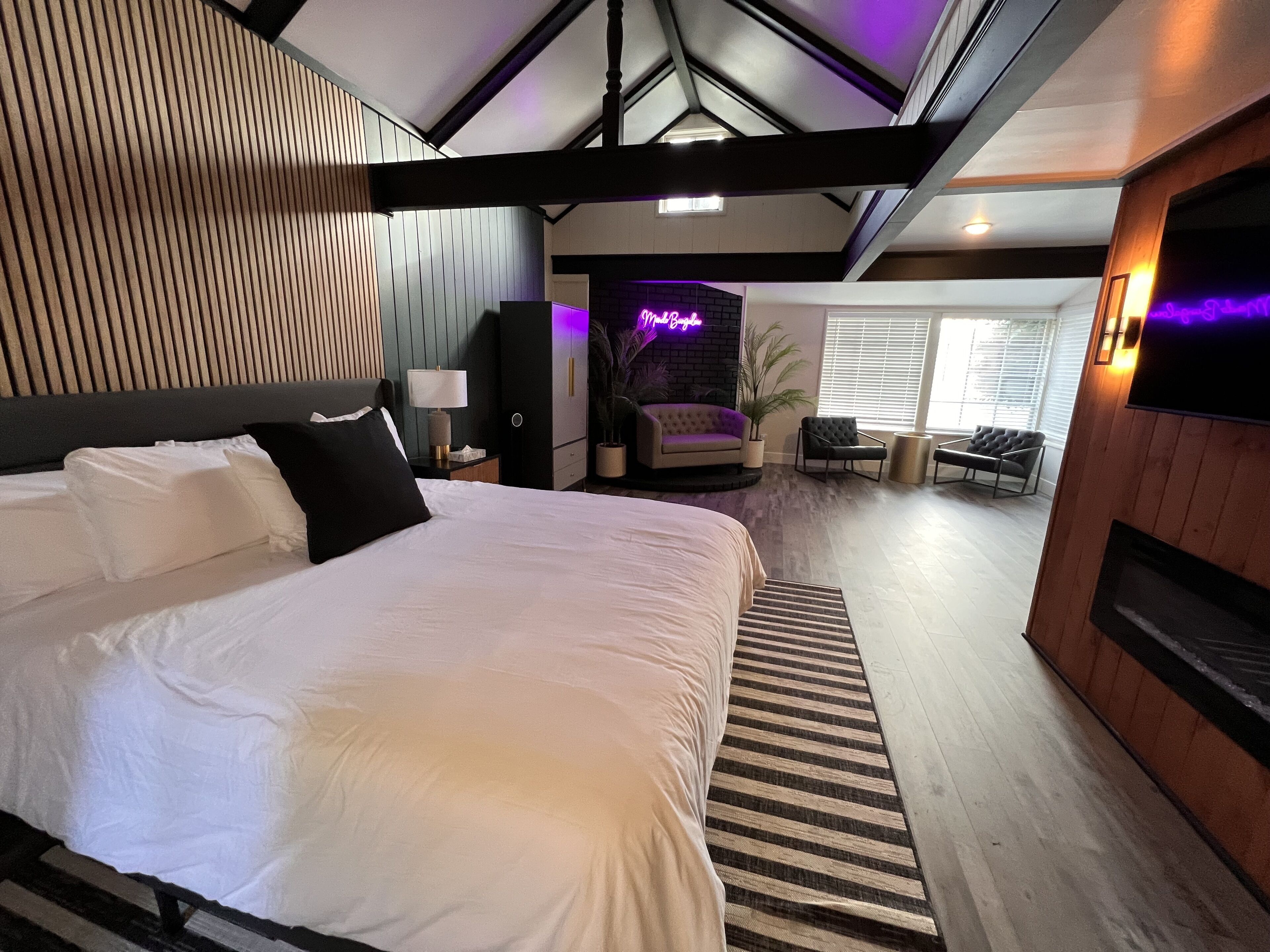 Una habitación de hotel moderna con una cama grande, un televisor de pantalla plana, un letrero de neón y una zona de estar acogedora.