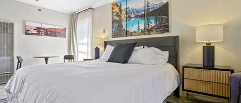 Habitación de hotel con una cama grande, una mesita de noche con lámpara, una mesita pequeña y una silla. Hay dos cuadros en la pared.
