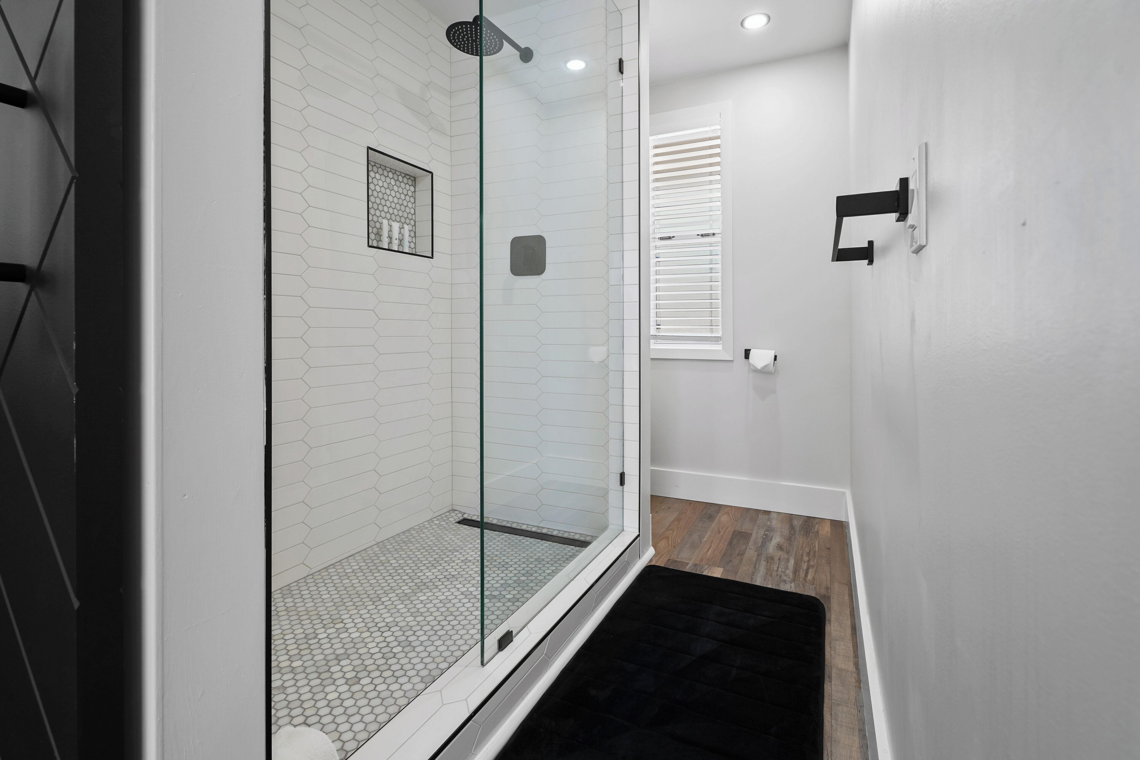 A modern bathroom with a glass shower enclosure, white tiled walls, and a black rug.