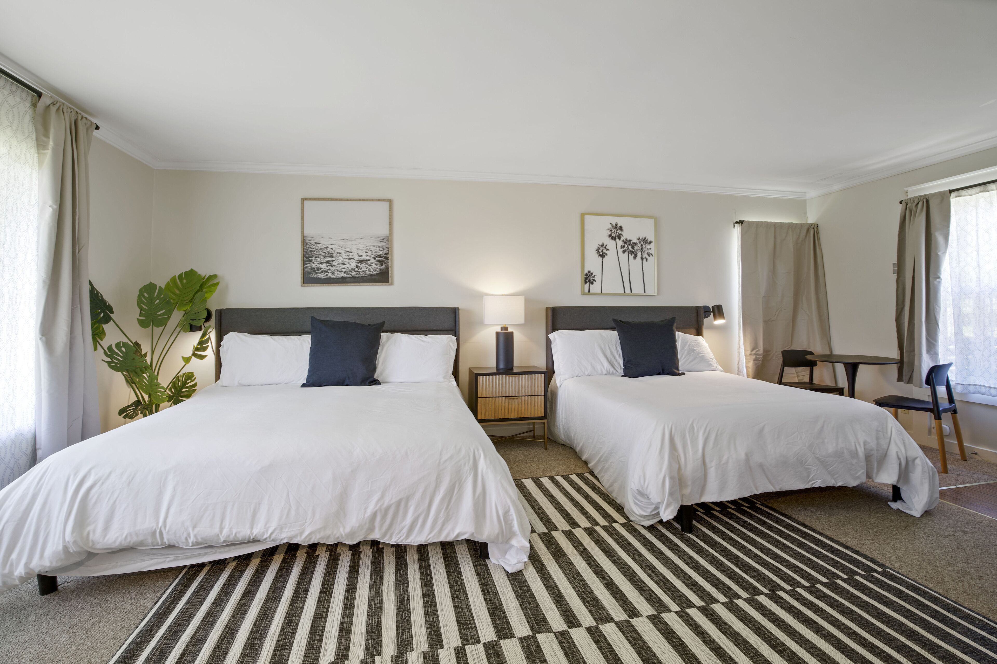 A hotel room with two beds, a bedside table with a lamp, a large plant, and artwork on the wall.