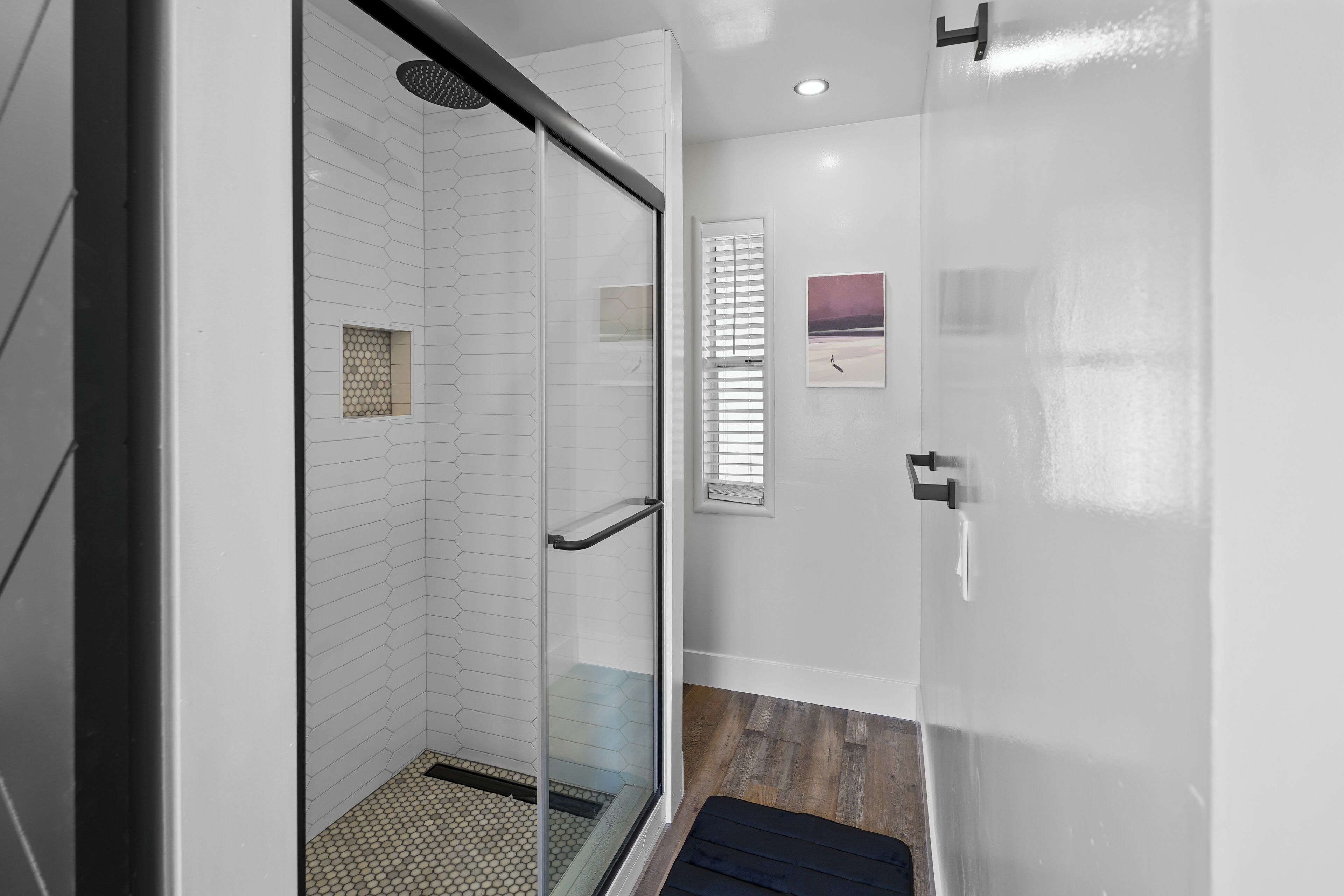 Une salle de bain moderne avec une douche à parois de verre, un plancher en bois et une petite fenêtre avec des stores.
