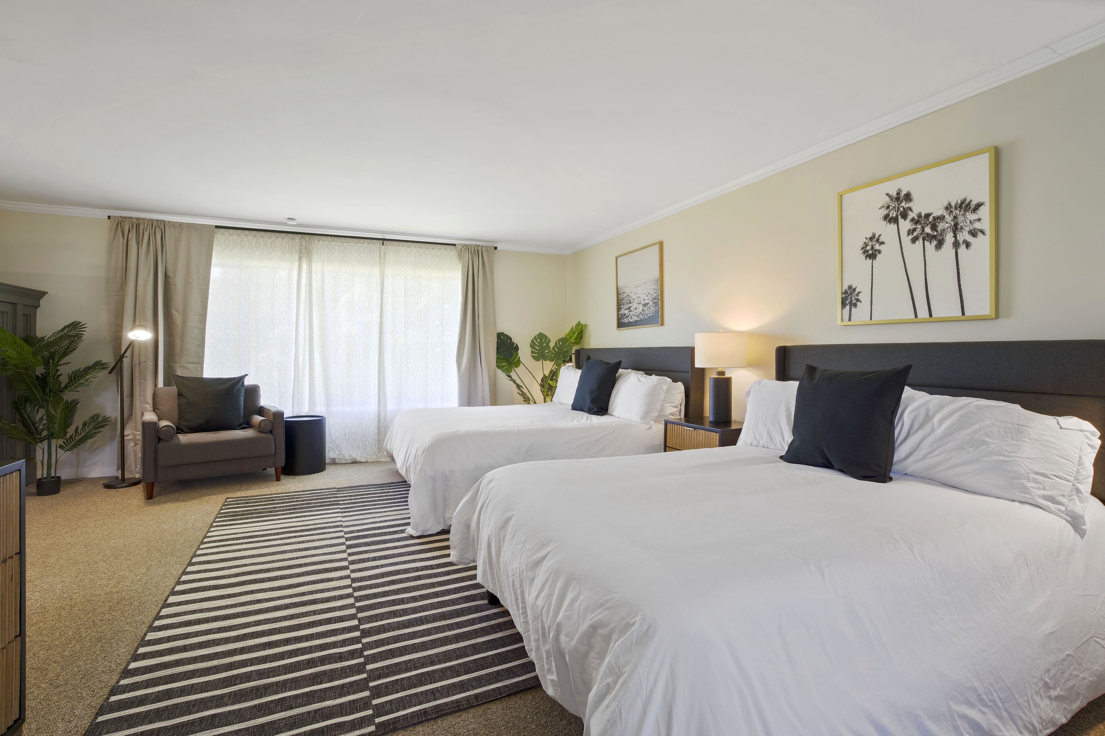 Quarto de hotel com duas camas, um sofá, uma janela grande com cortinas, um vaso de planta e um quadro na parede.