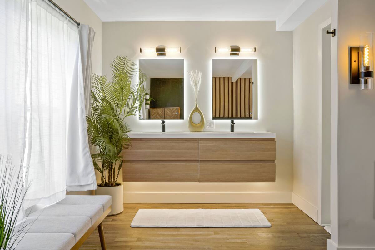 Uma casa de banho moderna com toucador de madeira, dois espelhos, uma planta em vaso e um banco.