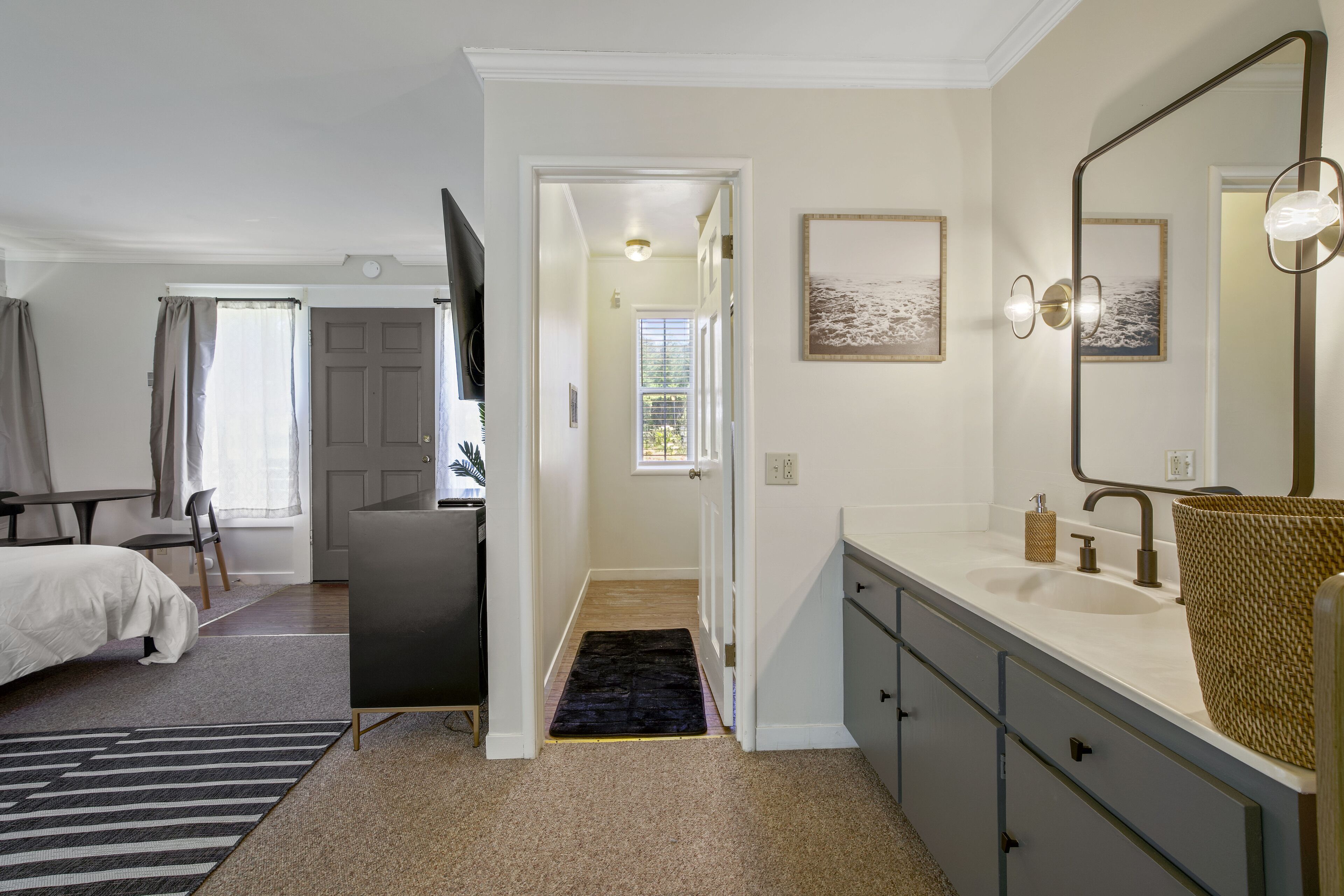 A modern bedroom with a bed, bedside table, and a large mirror. A hallway leads to a bathroom with a sink and a towel rack.