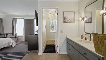 A modern bedroom with a bed, bedside table, and a large mirror. A hallway leads to a bathroom with a sink and a towel rack.