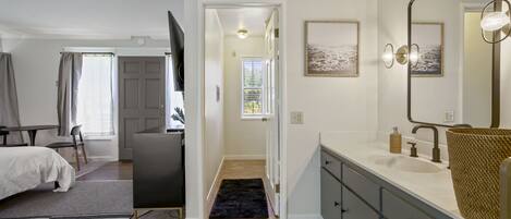 A modern bedroom with a bed, bedside table, and a large mirror. A hallway leads to a bathroom with a sink and a towel rack.