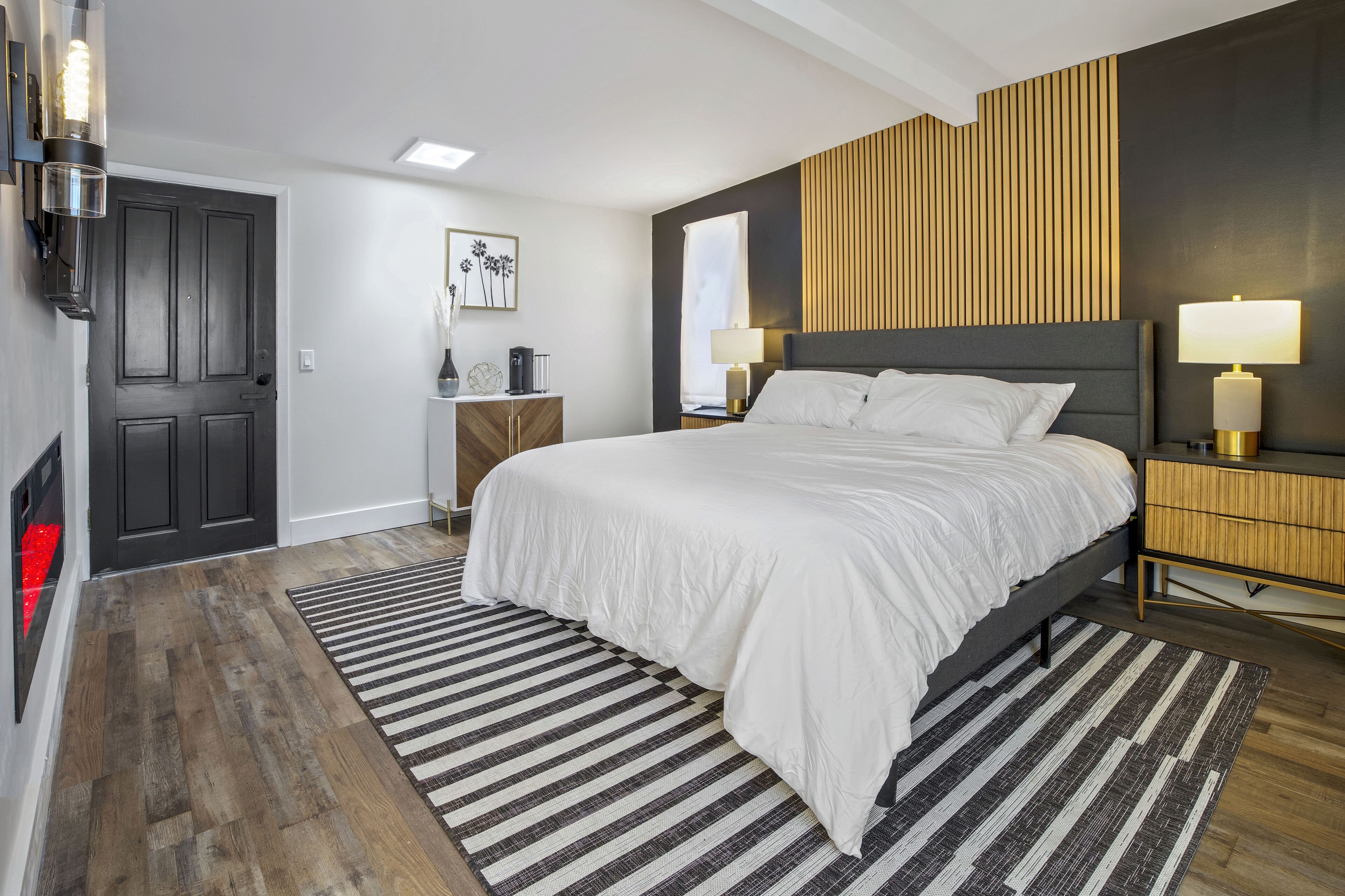 A modern bedroom with a large bed, a wooden headboard, a nightstand with a lamp, and a striped rug.