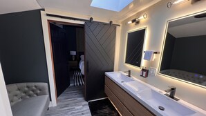 A modern bathroom with a large mirror, dual sinks, and a sliding door leading to a bedroom. - Mendo Bungalow (Mendocino)