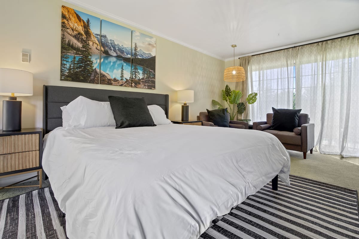 Quarto de hotel com uma cama grande, uma mesa de cabeceira, uma luminária, uma cadeira e uma janela ampla com cortinas.