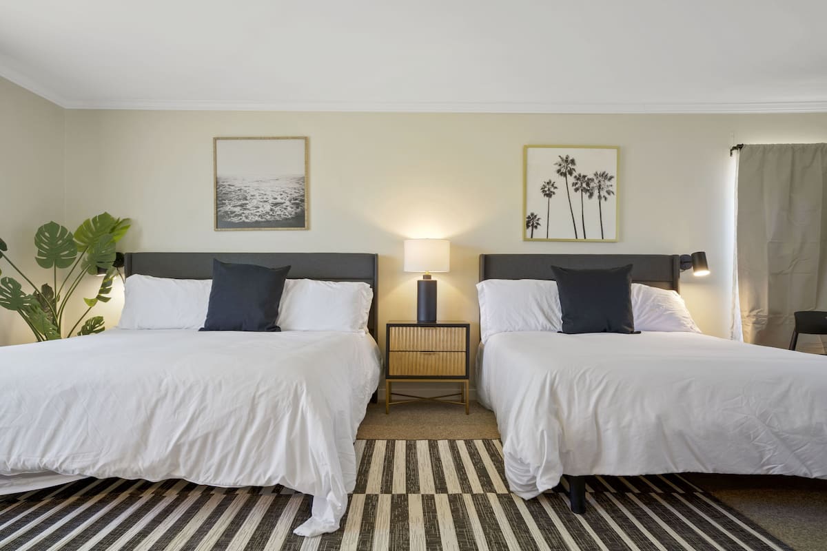 Quarto de hotel com duas camas, uma mesa de cabeceira, uma luminária e duas fotos emolduradas na parede.