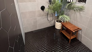 A modern bathroom with hexagonal patterned floor tiles, a wooden bench, a potted plant, a showerhead, and a small shelf with toiletries. - Mendo Bungalow (Mendocino)