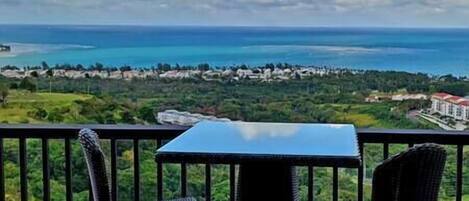 Un balcón con una mesa y dos sillas, con vistas a un paisaje costero y al mar, donde se divisa una orilla lejana.