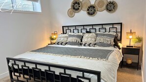 A bed with a black metal frame, white bedding, and decorative pillows. There are two bedside tables with lamps and a wall-mounted decorative item above the bed. - BoHo Modern Retreat w/King|Fireplace|Disney+ (Edmonton)