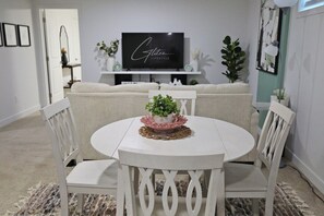Ein Essbereich mit einem runden Tisch, weißen Stühlen und einem dekorativen Mittelstück. Ein Flachbildfernseher ist an der Wand über dem Tisch angebracht.