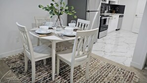 Un comedor con una mesa blanca dispuesta para cuatro personas, rodeada de sillas blancas. Una planta pequeña está sobre la mesa, y se puede ver una cocina al fondo con armarios y electrodomésticos blancos.