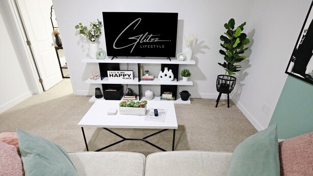 Una sala de estar con un sofá blanco, una mesa de centro, un televisor sobre un mueble y un estante con objetos decorativos.