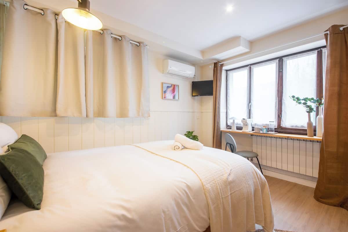 A hotel room with a bed, a desk with a chair, a window with curtains, an air conditioning unit, and a television mounted on the wall.