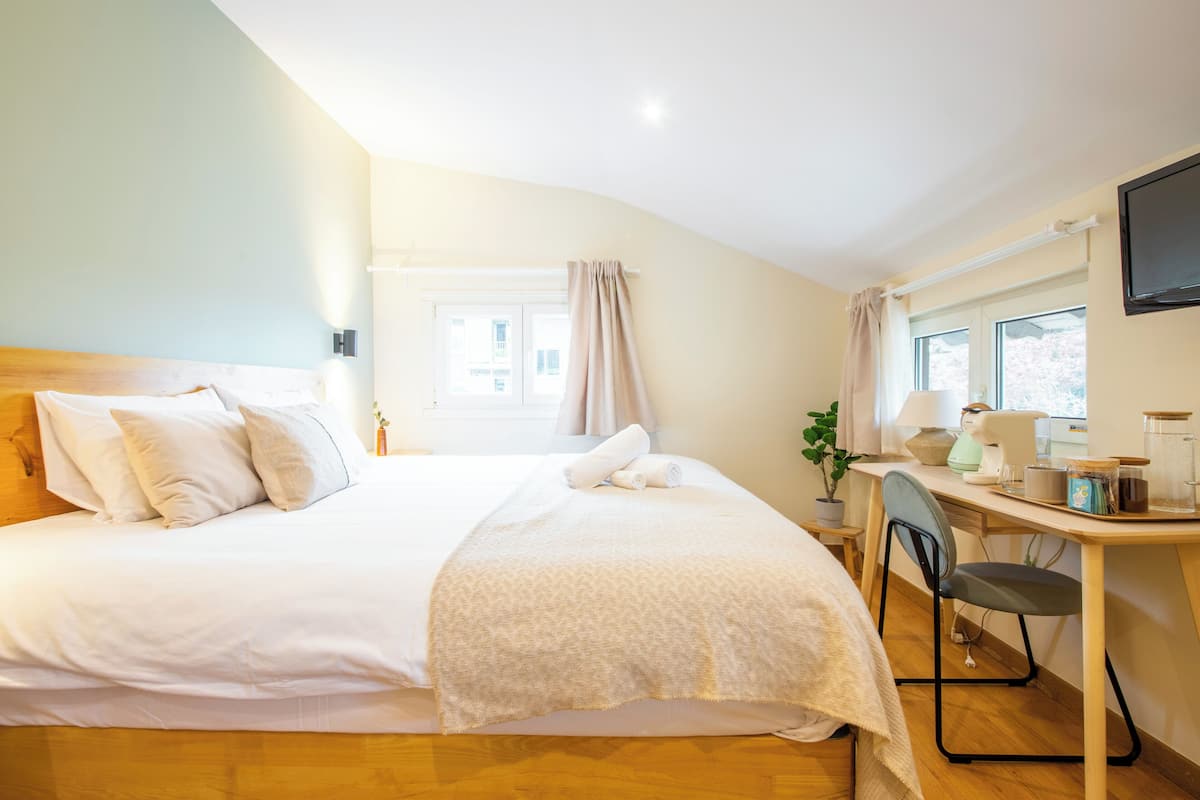 A neatly made bed with white linens, a desk with a lamp, and a window with curtains.