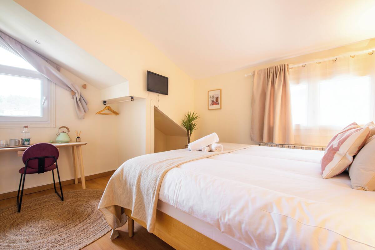 A bedroom with a bed, a desk, a chair, a TV, and a window with curtains.