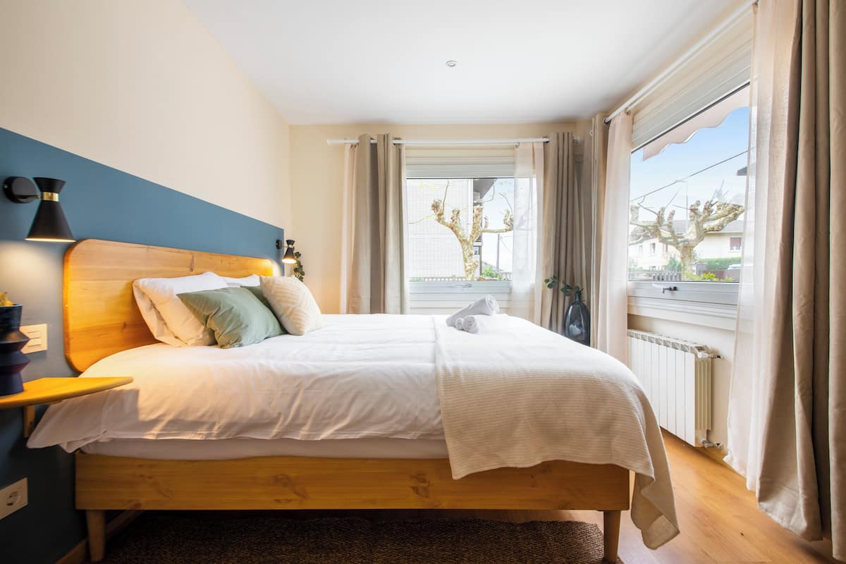 A bedroom with a wooden bed, white bedding, a nightstand with a lamp, a view of the outside through a window with curtains, and a radiator under the window.