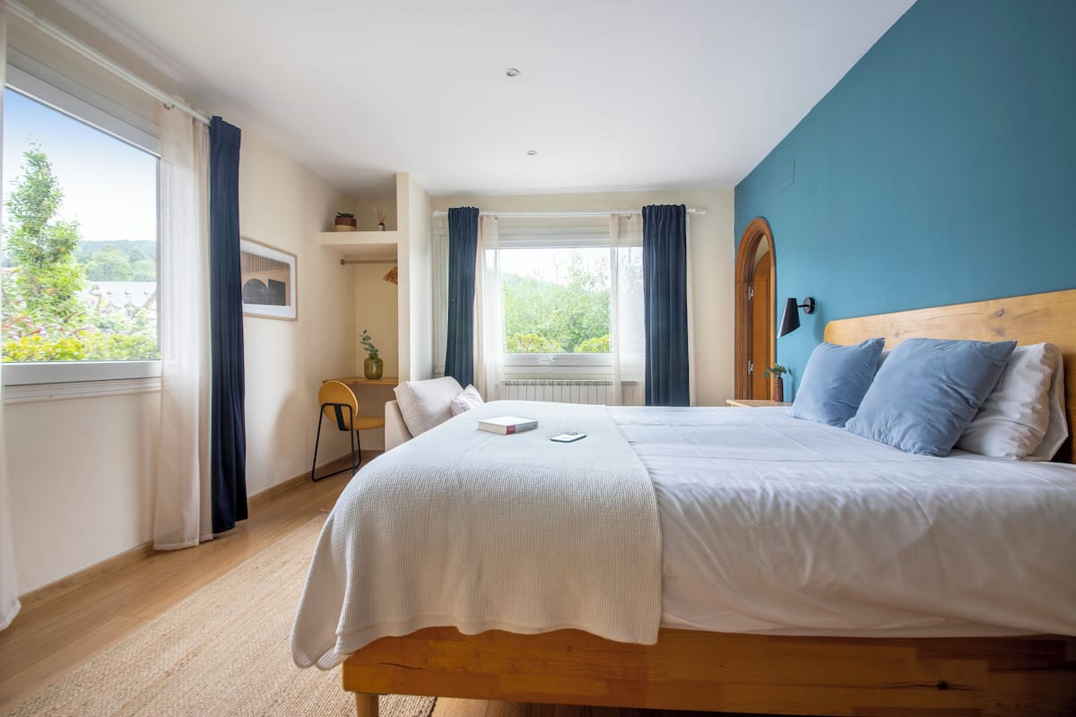 A bedroom with a large bed, a view of greenery outside, and a window with curtains.