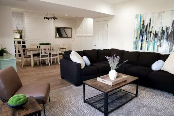 A modern living room with a black sofa, a wooden coffee table, and a dining area in the background.