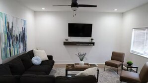 A modern living room with a black sofa, a brown armchair, a coffee table, and a flat-screen TV mounted on the wall. - Spacious, Updated 4-bedroom Home near Short North Arts / Entertainment District (Columbus)