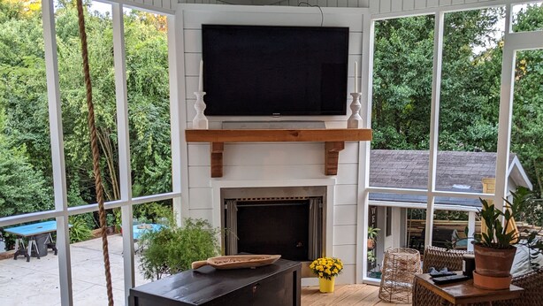 A covered outdoor living area with a fireplace, a television, wicker furniture, and a view of the surrounding greenery. - Master's Tournament Rental (Augusta)