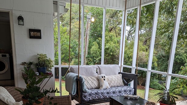A covered porch with a swing chair, a sofa, and a coffee table. There are potted plants and a washing machine visible. - Charming Country Cottage Near Augusta National (Augusta)
