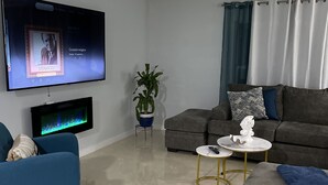 A modern living room with a flat-screen TV, a grey sofa, a blue armchair, a small round coffee table, and a potted plant.