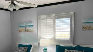 A bedroom with two beds, a ceiling fan, a window with blinds, and artwork on the walls.