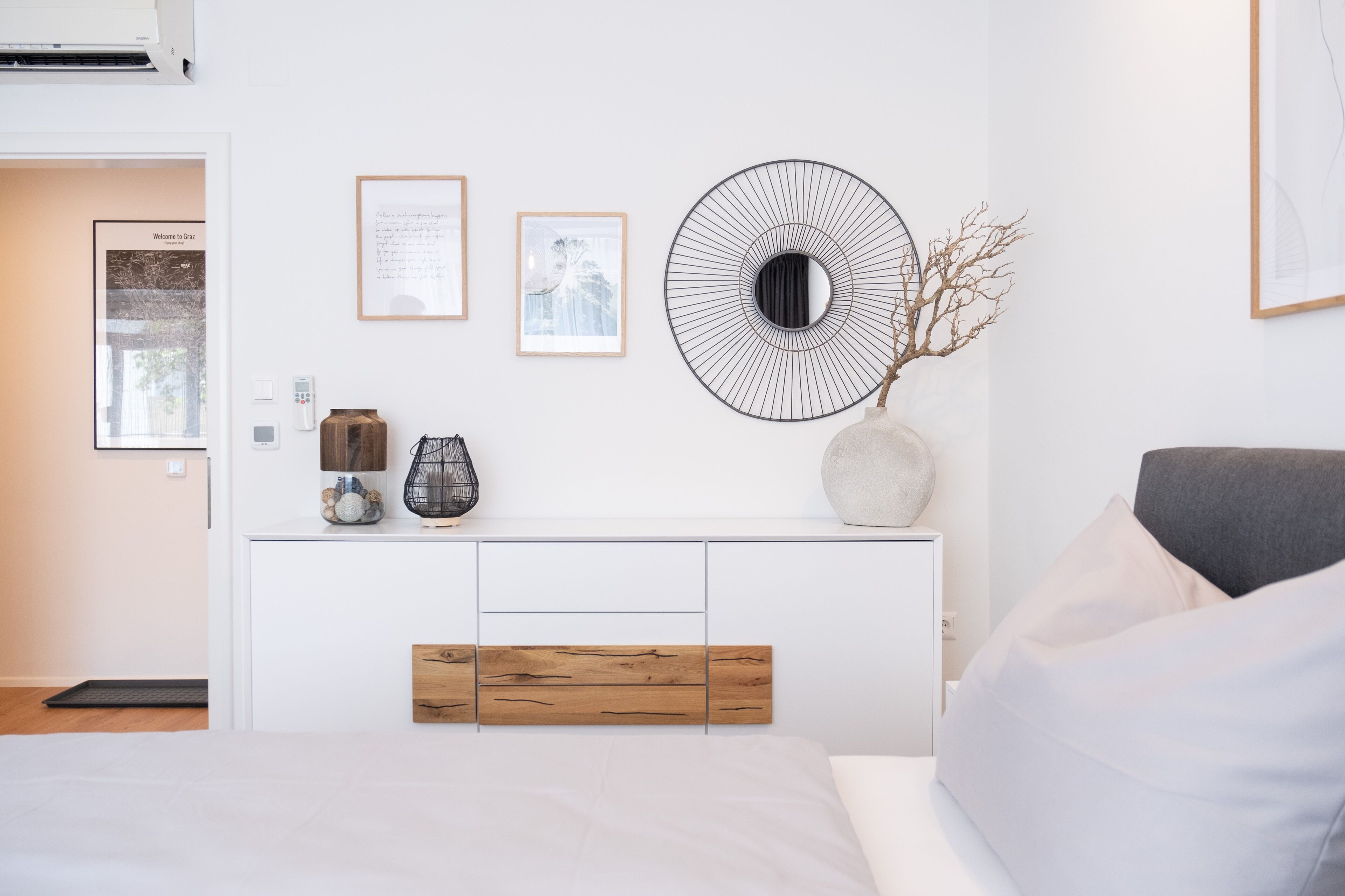 Ein modernes Schlafzimmer mit einem weißen Bett, einem grauen Sofa, einem Holzkonsolentisch mit Dekoration und Wandbildern.