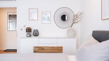 Ein modernes Schlafzimmer mit einem weißen Bett, einem grauen Sofa, einem Holzkonsolentisch mit Dekoration und Wandbildern.