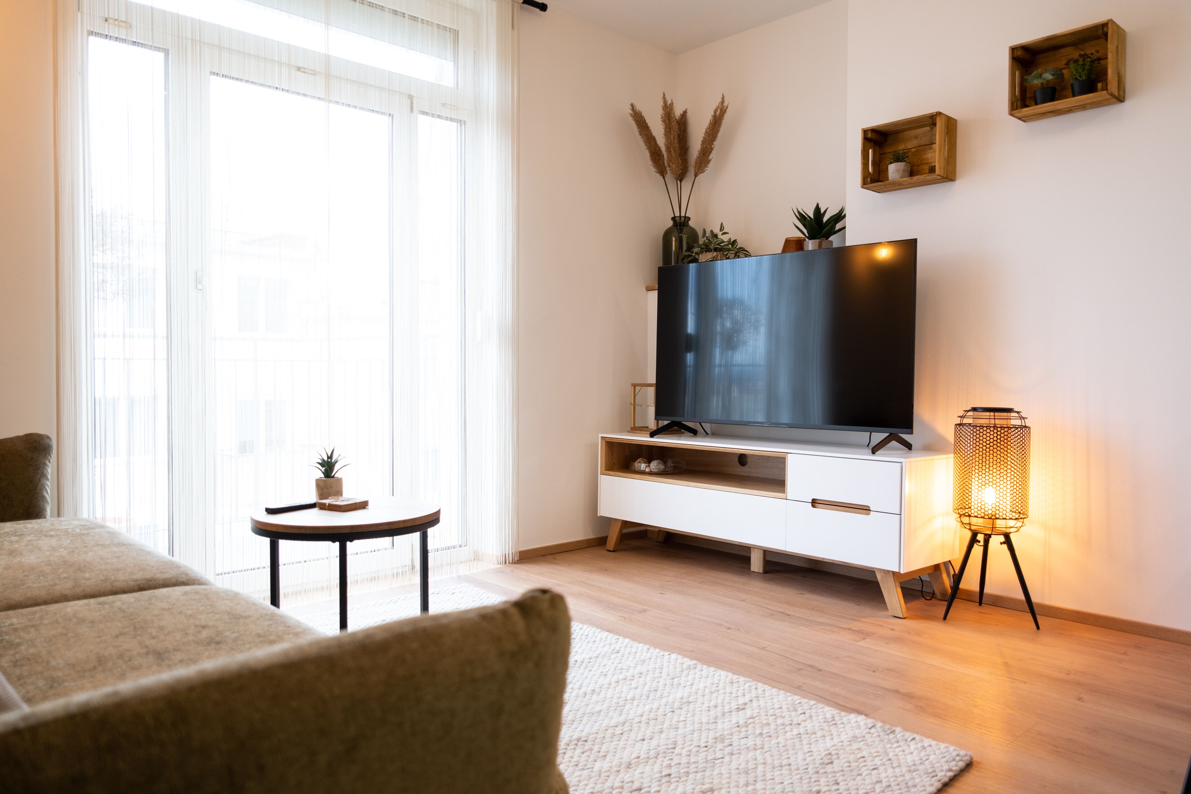 Una sala de estar moderna con un televisor de pantalla plana, una mesita auxiliar con una planta, una lámpara de pie y un ventanal con cortinas translúcidas.