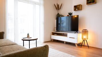 Una sala de estar moderna con un televisor de pantalla plana, una mesita auxiliar con una planta, una lámpara de pie y un ventanal con cortinas translúcidas.
