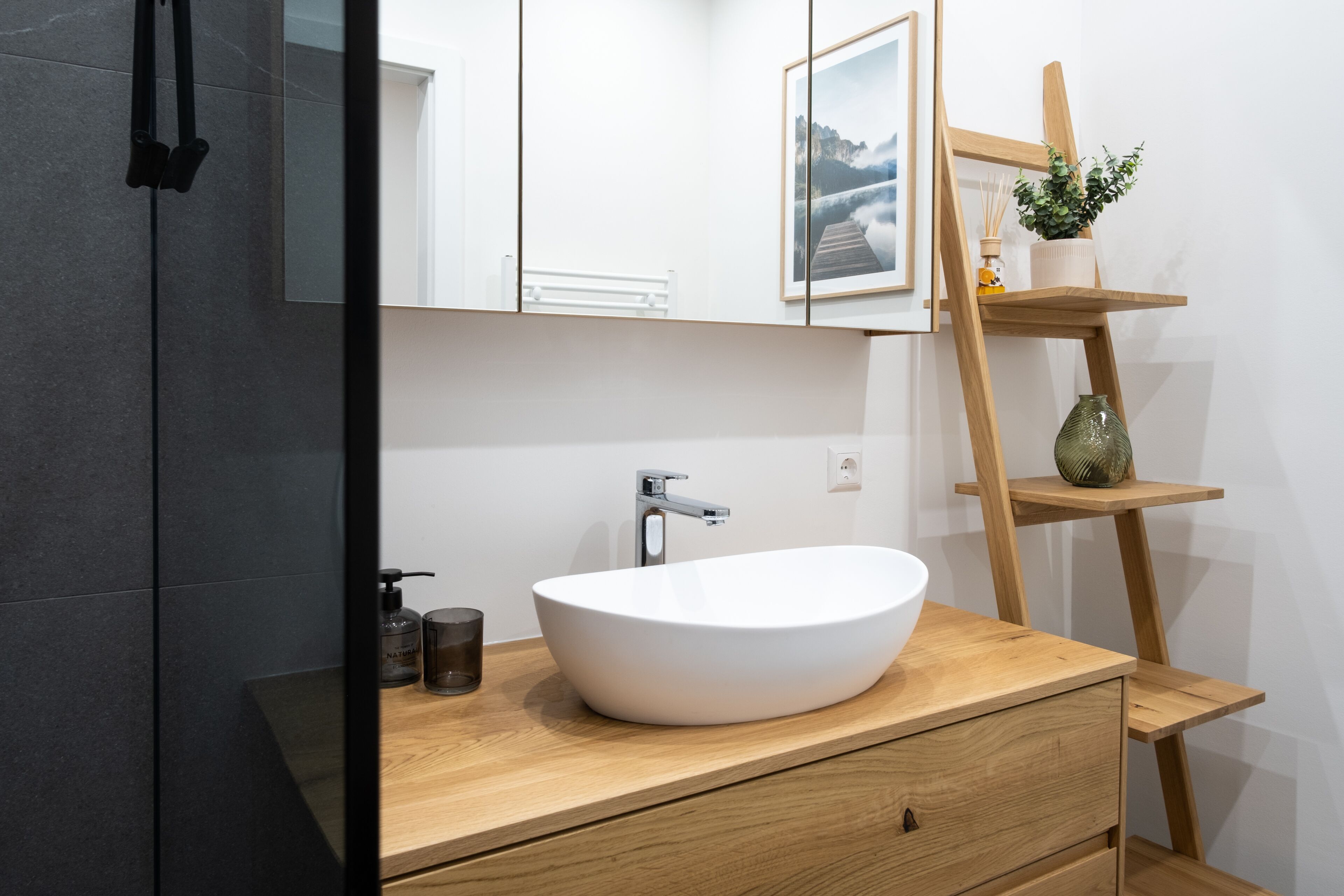 Un baño moderno con un lavamanos blanco de diseño, tocador de madera, un espejo y un estante escalonado de madera con una planta y un florero.