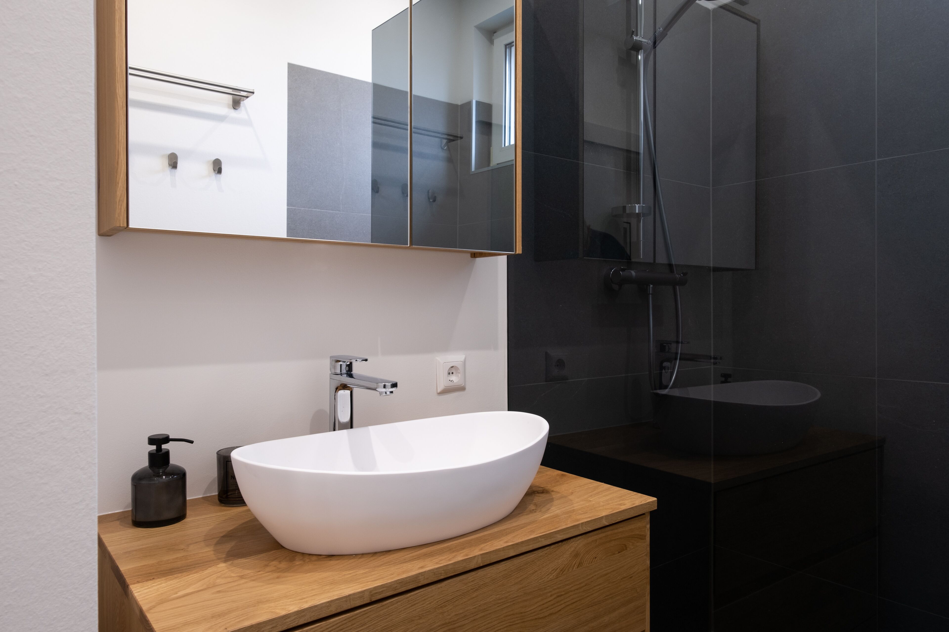 A modern bathroom with a large white vessel sink, a wooden vanity, a mirror, and a glass-enclosed shower area.