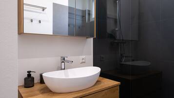 Un baño moderno con un lavamanos blanco grande tipo recipiente, un mueble de tocador de madera, un espejo y una zona de ducha con mampara de vidrio.