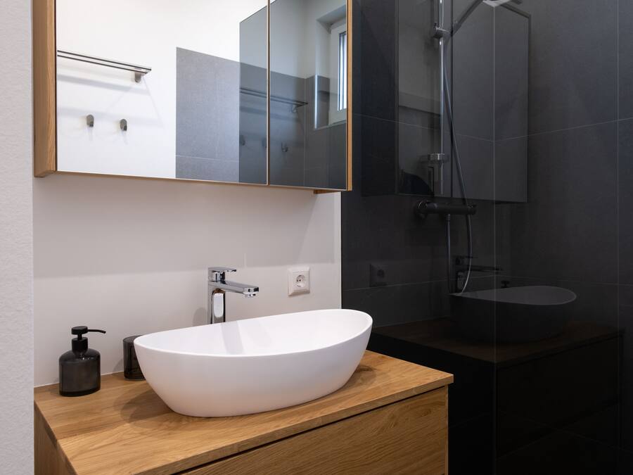 Un baño moderno con un lavamanos blanco grande tipo recipiente, un mueble de tocador de madera, un espejo y una zona de ducha con mampara de vidrio.