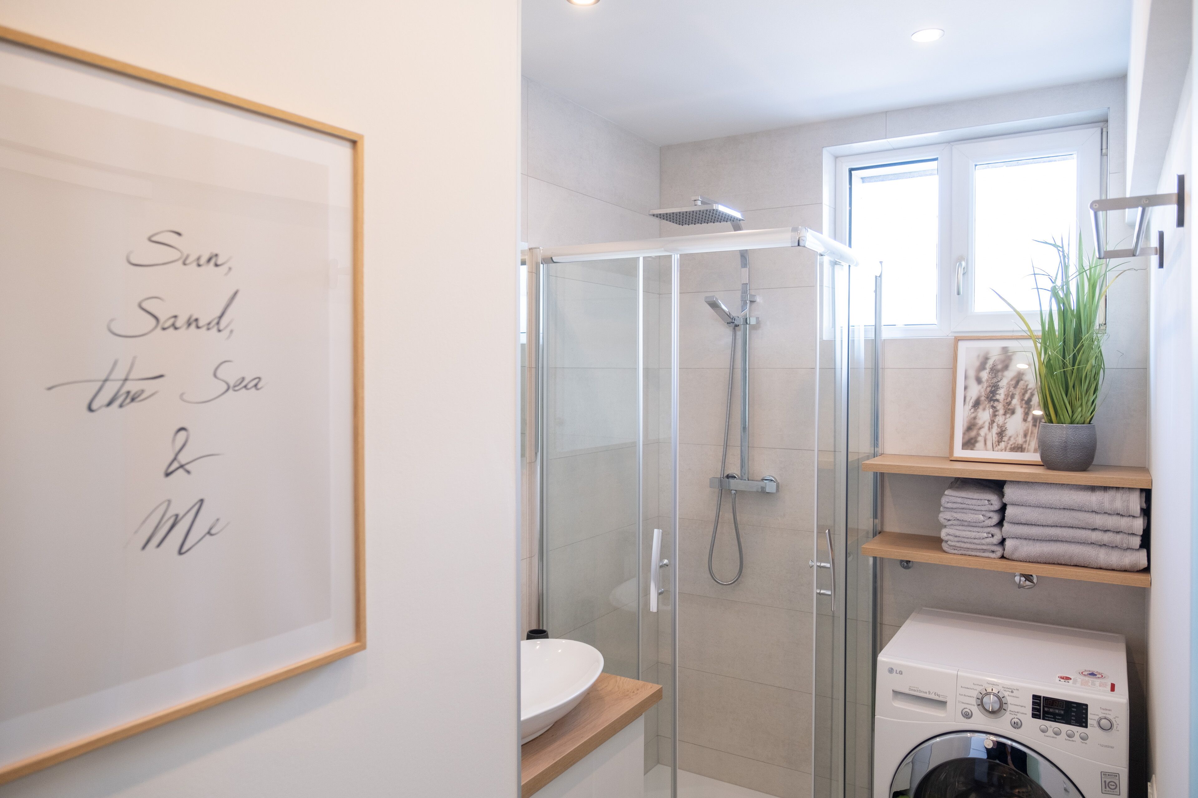 A modern bathroom with a glass shower enclosure, a framed quote on the wall, a washing machine, and a window providing natural light.